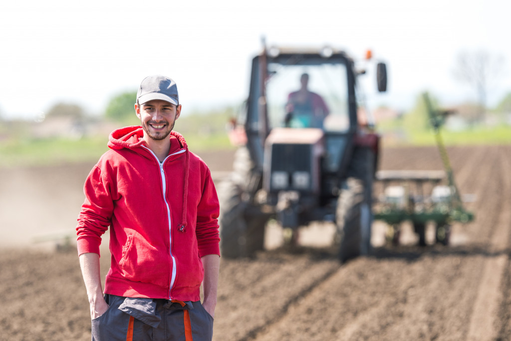 a smiling man in a red hoodie standing in a field being plowed by a tractor in the background